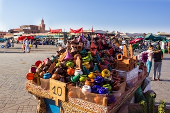 A vibrant market scene features a stall filled with colorful ceramic tagines and traditional pottery. Various stalls and people are visible in the bustling background, with a historic tower and buildings providing a scenic backdrop. Red tents and awnings add to the lively atmosphere.