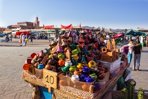 A vibrant market scene features a stall filled with colorful ceramic tagines and traditional pottery. Various stalls and people are visible in the bustling background, with a historic tower and buildings providing a scenic backdrop. Red tents and awnings add to the lively atmosphere.