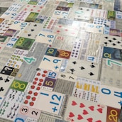 Numerous playing cards and educational number cards are laid out in a patchwork pattern on a surface. The cards are of various styles and designs, including traditional playing cards, cards featuring mathematical equations, and cards with different numerical values and graphics, such as apples, dots, and shapes.