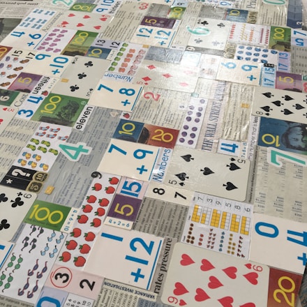 Numerous playing cards and educational number cards are laid out in a patchwork pattern on a surface. The cards are of various styles and designs, including traditional playing cards, cards featuring mathematical equations, and cards with different numerical values and graphics, such as apples, dots, and shapes.