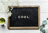 A small blackboard with a wooden frame displays the word 'COOL' in white letters. Surrounding the board on a white wooden surface are delicate white flowers and greenery. A cup of green matcha tea is placed to the left.