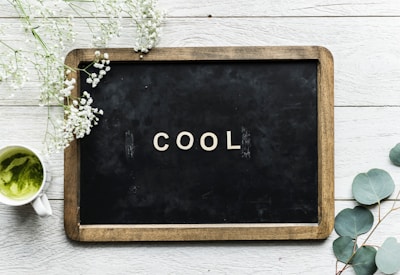 A small blackboard with a wooden frame displays the word 'COOL' in white letters. Surrounding the board on a white wooden surface are delicate white flowers and greenery. A cup of green matcha tea is placed to the left.