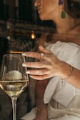 A woman wearing elegant jewelry raises a glass of white wine, with reflections from the glass creating an intriguing play of light. Her nails are polished, and she wears a strapless white outfit with bold earrings. The scene appears to be indoors, possibly in a sophisticated setting.