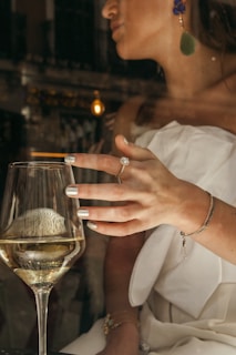 A woman wearing elegant jewelry raises a glass of white wine, with reflections from the glass creating an intriguing play of light. Her nails are polished, and she wears a strapless white outfit with bold earrings. The scene appears to be indoors, possibly in a sophisticated setting.