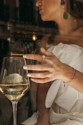 A woman wearing elegant jewelry raises a glass of white wine, with reflections from the glass creating an intriguing play of light. Her nails are polished, and she wears a strapless white outfit with bold earrings. The scene appears to be indoors, possibly in a sophisticated setting.