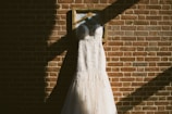 A wedding dress is elegantly displayed on a wooden hanger, hanging against a textured brick wall. The sunlight creates shadows and highlights, adding depth and contrast to the scene. The dress features intricate lace details and a flowing train.