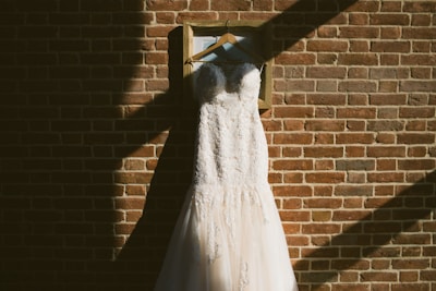 A wedding dress is elegantly displayed on a wooden hanger, hanging against a textured brick wall. The sunlight creates shadows and highlights, adding depth and contrast to the scene. The dress features intricate lace details and a flowing train.