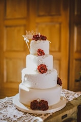 A three-tiered white wedding cake adorned with intricate icing patterns and decorated with red roses. The cake is embellished with strings of pearls and is set on a lace-covered table. Soft, warm lighting casts a subtle glow on the cake, highlighting its elegant design. A small decorative sign with initials is placed on the top tier.