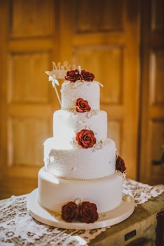 A three-tiered white wedding cake adorned with intricate icing patterns and decorated with red roses. The cake is embellished with strings of pearls and is set on a lace-covered table. Soft, warm lighting casts a subtle glow on the cake, highlighting its elegant design. A small decorative sign with initials is placed on the top tier.