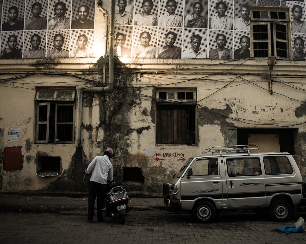 man in white shirt beside black motor scooter looking up at sketch portraits hanged on building wall