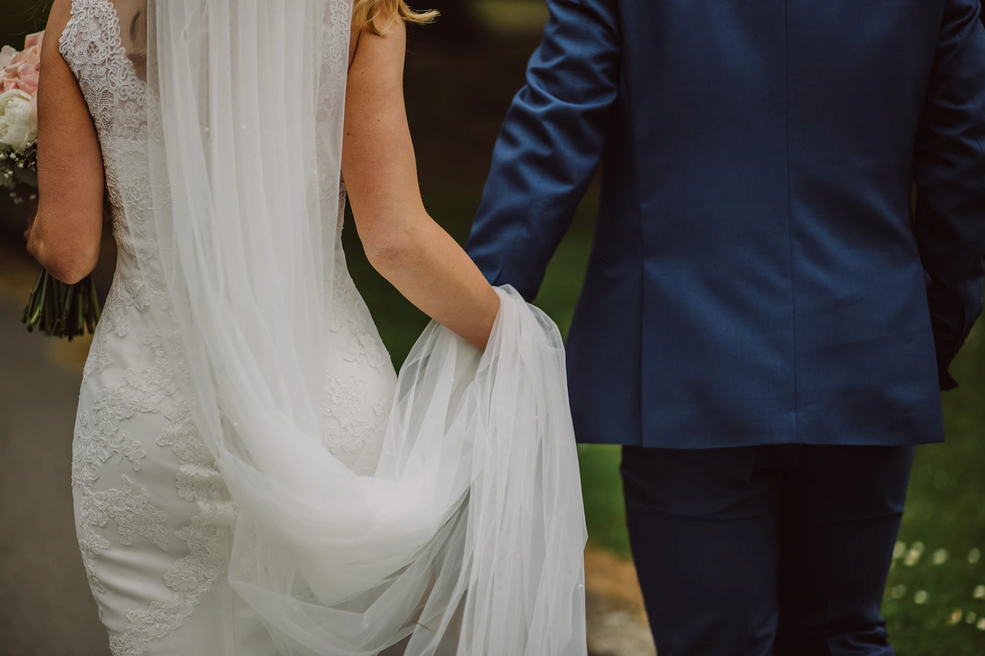 Bride in white lace wedding gown and groom in navy suit walking hand in hand from behind with soft golden light