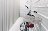 A bicycle is parked in a narrow, enclosed space between two corrugated metal walls. The focus is on the back of the bicycle, which features a large wooden crate attached to its rear. The surroundings and the bike are both in shades of gray and white, giving a minimalist and industrial feel.