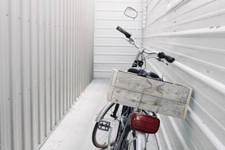 A bicycle is parked in a narrow, enclosed space between two corrugated metal walls. The focus is on the back of the bicycle, which features a large wooden crate attached to its rear. The surroundings and the bike are both in shades of gray and white, giving a minimalist and industrial feel.
