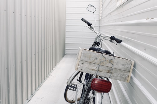 A bicycle is parked in a narrow, enclosed space between two corrugated metal walls. The focus is on the back of the bicycle, which features a large wooden crate attached to its rear. The surroundings and the bike are both in shades of gray and white, giving a minimalist and industrial feel.