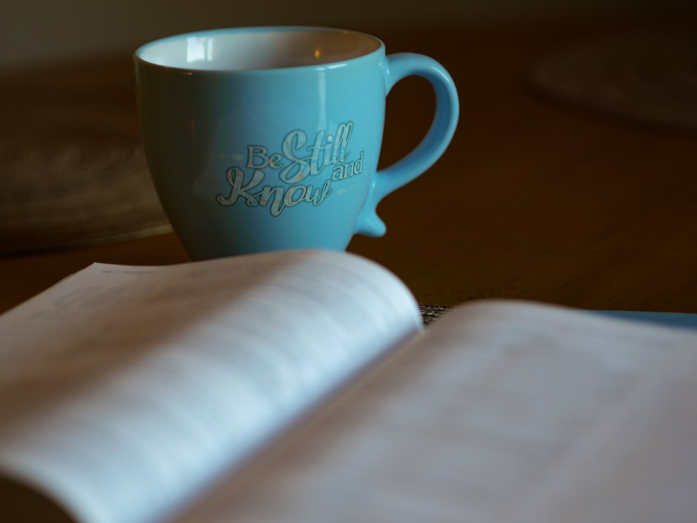 Flachfokusfotografie eines aufgeschlagenen Buches neben einer blauen Keramiktasse