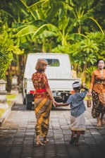 A woman and a child in traditional attire are holding hands and walking on a paved path. The child is wearing a traditional headdress, and a white truck is parked ahead. Surrounding them are lush green tropical plants, creating a vibrant and calm atmosphere.