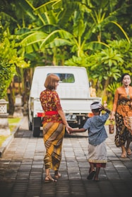 A woman and a child in traditional attire are holding hands and walking on a paved path. The child is wearing a traditional headdress, and a white truck is parked ahead. Surrounding them are lush green tropical plants, creating a vibrant and calm atmosphere.