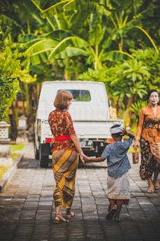 A woman and a child in traditional attire are holding hands and walking on a paved path. The child is wearing a traditional headdress, and a white truck is parked ahead. Surrounding them are lush green tropical plants, creating a vibrant and calm atmosphere.