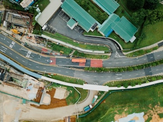 An aerial view of a roadway complex surrounded by greenery and buildings with green roofs. The roads are winding and there are multiple lanes with a few vehicles, including a truck and a car. Pedestrian paths are visible, along with a construction site area nearby. The landscape is a mix of structured urban elements and natural green spaces.