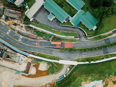 An aerial view of a roadway complex surrounded by greenery and buildings with green roofs. The roads are winding and there are multiple lanes with a few vehicles, including a truck and a car. Pedestrian paths are visible, along with a construction site area nearby. The landscape is a mix of structured urban elements and natural green spaces.