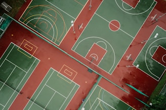 Aerial view of outdoor sports courts displaying multiple basketball and tennis courts positioned side by side. The courts are painted with green playing surfaces and red surrounding areas, with white and yellow boundary lines. The basketball courts feature hoops and painted three-point arcs and key areas, while the tennis courts include service lines and baselines.