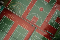 Aerial view of outdoor sports courts displaying multiple basketball and tennis courts positioned side by side. The courts are painted with green playing surfaces and red surrounding areas, with white and yellow boundary lines. The basketball courts feature hoops and painted three-point arcs and key areas, while the tennis courts include service lines and baselines.