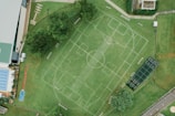 An aerial view of a sports complex featuring multiple sports fields drawn on green grass. The layout includes soccer and tennis courts, with pathways surrounding the area. Trees and surrounding buildings are visible, along with a swimming pool section on the left side.