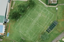 An aerial view of a sports complex featuring multiple sports fields drawn on green grass. The layout includes soccer and tennis courts, with pathways surrounding the area. Trees and surrounding buildings are visible, along with a swimming pool section on the left side.