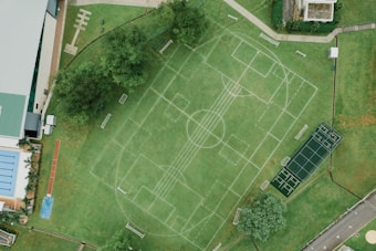 An aerial view of a sports complex featuring multiple sports fields drawn on green grass. The layout includes soccer and tennis courts, with pathways surrounding the area. Trees and surrounding buildings are visible, along with a swimming pool section on the left side.