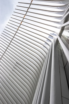 A modern architectural structure characterized by a series of white, curving, elongated ribs that create a dynamic and striking visual effect. The ribs rise upward and curve inwards, forming a grid-like pattern with gaps in between, allowing light to pass through.