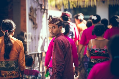 A group of people in vibrant traditional clothing are walking together outdoors. One man in a maroon shirt and headpiece turns to look back, making eye contact with the viewer. The scene is lively, highlighting cultural attire with bright colors and patterns, set against a sunny backdrop.