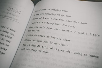 The image features an open book displaying text in two columns. The right column consists of a poem or song lyrics in English, expressing themes of longing and sadness. The left column contains text in Vietnamese. The page on the left also shows a timestamp, '01:34,' and a date, referring to Sunday, May 22, 2016.
