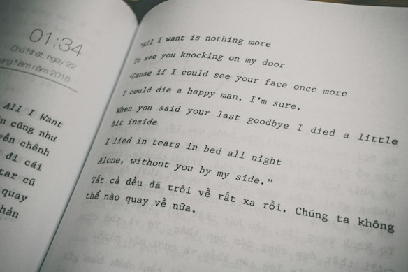 The image features an open book displaying text in two columns. The right column consists of a poem or song lyrics in English, expressing themes of longing and sadness. The left column contains text in Vietnamese. The page on the left also shows a timestamp, '01:34,' and a date, referring to Sunday, May 22, 2016.