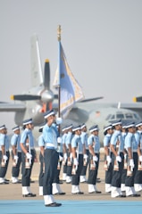 A group of uniformed individuals standing in formation, with one prominent person holding a flag. They are positioned on a tarmac, and there are military aircraft in the background. The uniforms are a combination of light blue shirts, dark pants, and white gloves, complemented by caps.