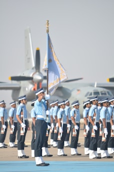 A group of uniformed individuals standing in formation, with one prominent person holding a flag. They are positioned on a tarmac, and there are military aircraft in the background. The uniforms are a combination of light blue shirts, dark pants, and white gloves, complemented by caps.