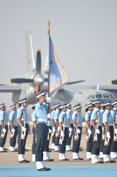 A group of uniformed individuals standing in formation, with one prominent person holding a flag. They are positioned on a tarmac, and there are military aircraft in the background. The uniforms are a combination of light blue shirts, dark pants, and white gloves, complemented by caps.
