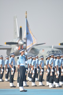 A group of uniformed individuals standing in formation, with one prominent person holding a flag. They are positioned on a tarmac, and there are military aircraft in the background. The uniforms are a combination of light blue shirts, dark pants, and white gloves, complemented by caps.