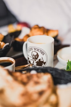A coffee cup with a logo from a café is positioned among a selection of pastries and breakfast items. The background appears slightly blurred, emphasizing the focal point of the cup in the foreground.