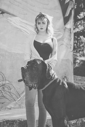 A woman with short hair stands confidently next to a large, black Great Dane. She wears a headband, a cardigan over a black top, and light-colored jeans. In the background, there is a large mural with abstract designs.
