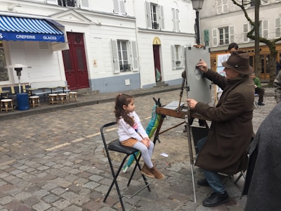 A street artist wearing a brown hat and coat is drawing a portrait on an easel, with a young girl sitting in front of him as the subject. They are in a cobblestone square with white buildings and a blue awning from a nearby café visible in the background. Some chairs are scattered around, and other people are present in the scene.
