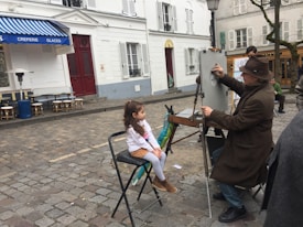 A street artist wearing a brown hat and coat is drawing a portrait on an easel, with a young girl sitting in front of him as the subject. They are in a cobblestone square with white buildings and a blue awning from a nearby café visible in the background. Some chairs are scattered around, and other people are present in the scene.