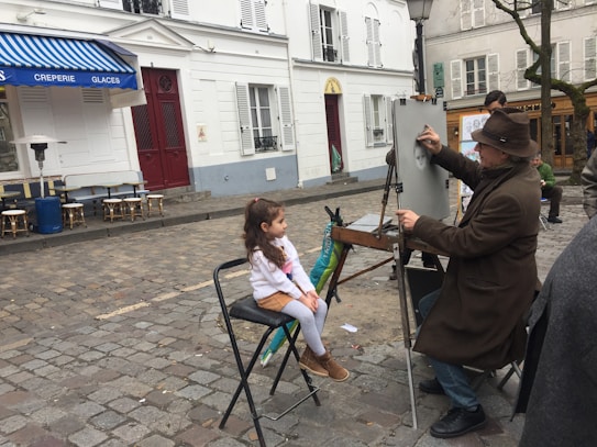 A street artist wearing a brown hat and coat is drawing a portrait on an easel, with a young girl sitting in front of him as the subject. They are in a cobblestone square with white buildings and a blue awning from a nearby caf&eacute; visible in the background. Some chairs are scattered around, and other people are present in the scene.