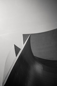 A monochromatic architectural photograph featuring sleek, curved metallic surfaces that are smoothly interlocking. The structure exhibits a seamless flow of geometric shapes, creating an elegant and modern aesthetic with a striking contrast between light and shadow.