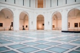 A spacious, elegant indoor hall with high ceilings and large archways. The floor is patterned with light-colored tiles, creating a modern geometric design. Artworks and sculptures are displayed along the gallery walls under soft lighting.