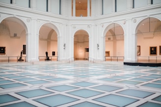 A spacious, elegant indoor hall with high ceilings and large archways. The floor is patterned with light-colored tiles, creating a modern geometric design. Artworks and sculptures are displayed along the gallery walls under soft lighting.