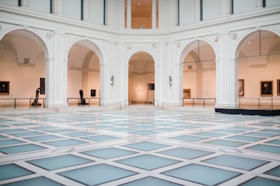 A spacious, elegant indoor hall with high ceilings and large archways. The floor is patterned with light-colored tiles, creating a modern geometric design. Artworks and sculptures are displayed along the gallery walls under soft lighting.