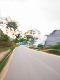 A blurred image of a winding road flanked by trees and a building. The motion blur suggests movement, creating a dynamic and fast-paced effect. The scene is set against a bright sky.
