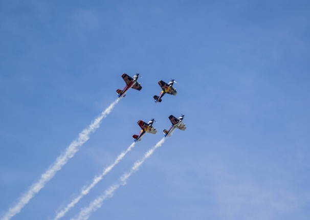 Four stunt airplanes fly in formation, leaving white smoke trails against a clear blue sky.
