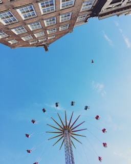 A vibrant amusement park ride spins high in the sky with several riders enjoying the thrill. The ride features colorful chairs suspended by chains, forming a circular pattern around a tall central tower. In the background, a clear blue sky dominates, with the corner of a building visible from below, adding an urban touch.