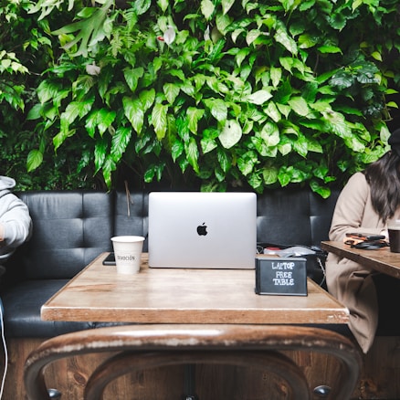 A laptop is placed on a wooden table in a cozy cafe setting, with a lush green leafy wall in the background. Two coffee cups are present, one with the branding 'Devoción'. A small sign reading 'Laptop Free Table' is on the table. Partially visible people are sitting on either side of the table, adding to the relaxed, informal ambiance.